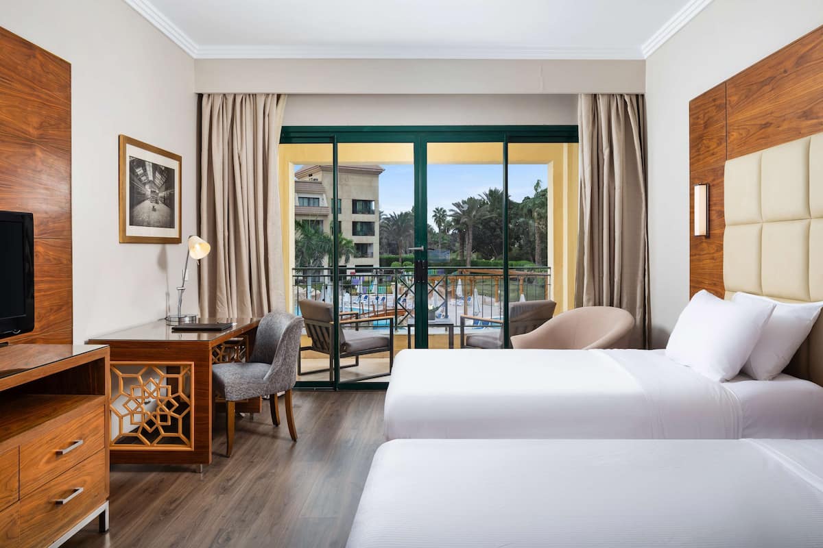 A hotel room with a bed, desk, chair, and a large window with a view of a pool and buildings.