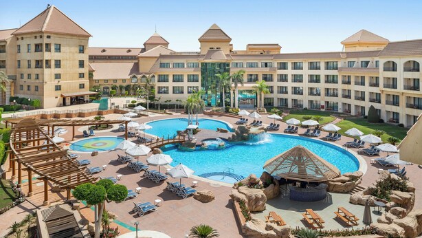 A hotel with a large central pool, multiple balconies, and a well-maintained garden area.