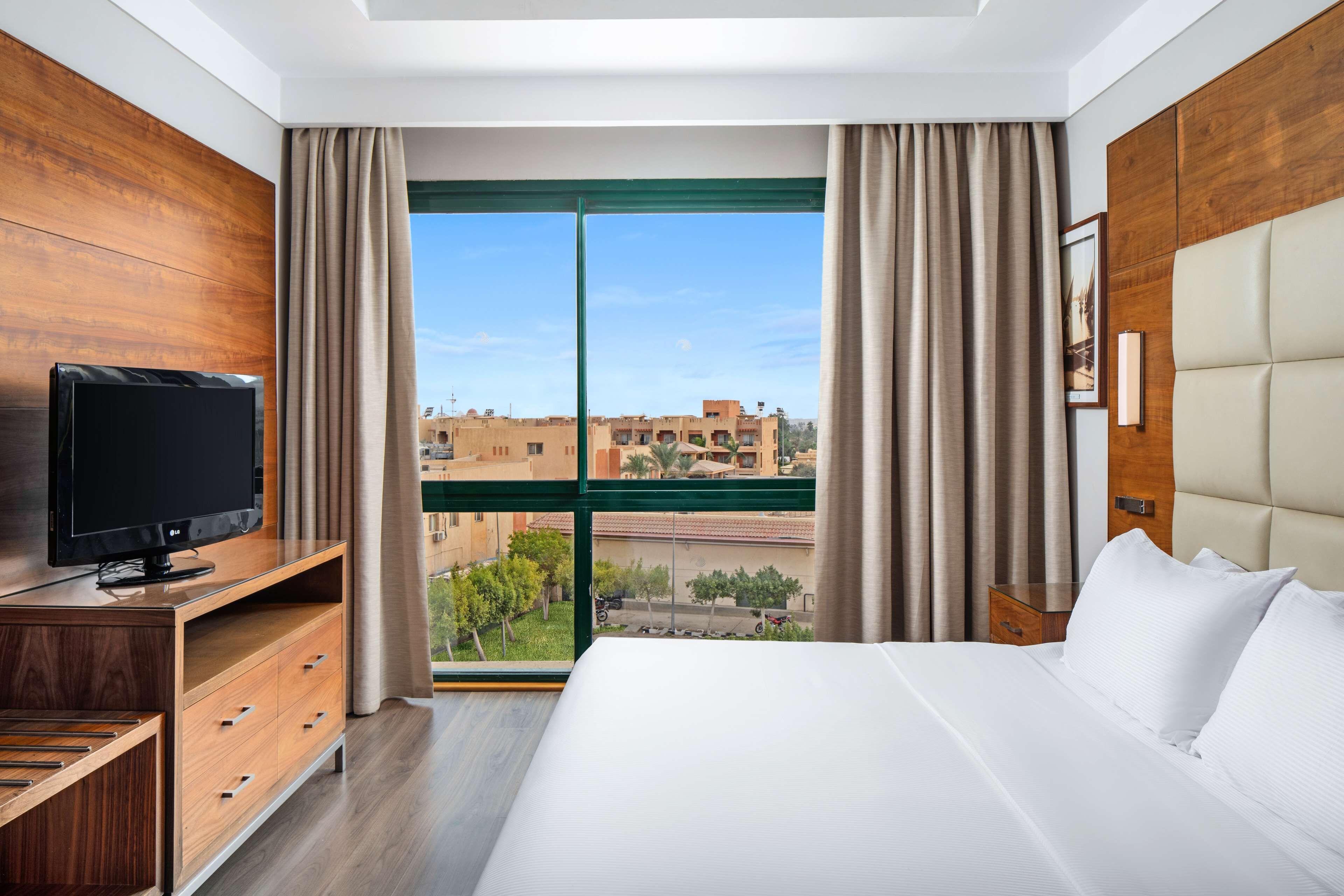 Ein Hotelzimmer mit einem großen Bett, Holzwandverkleidung, einem Fernseher auf einem Holzständer und Blick auf Gebäude durch ein großes Fenster.