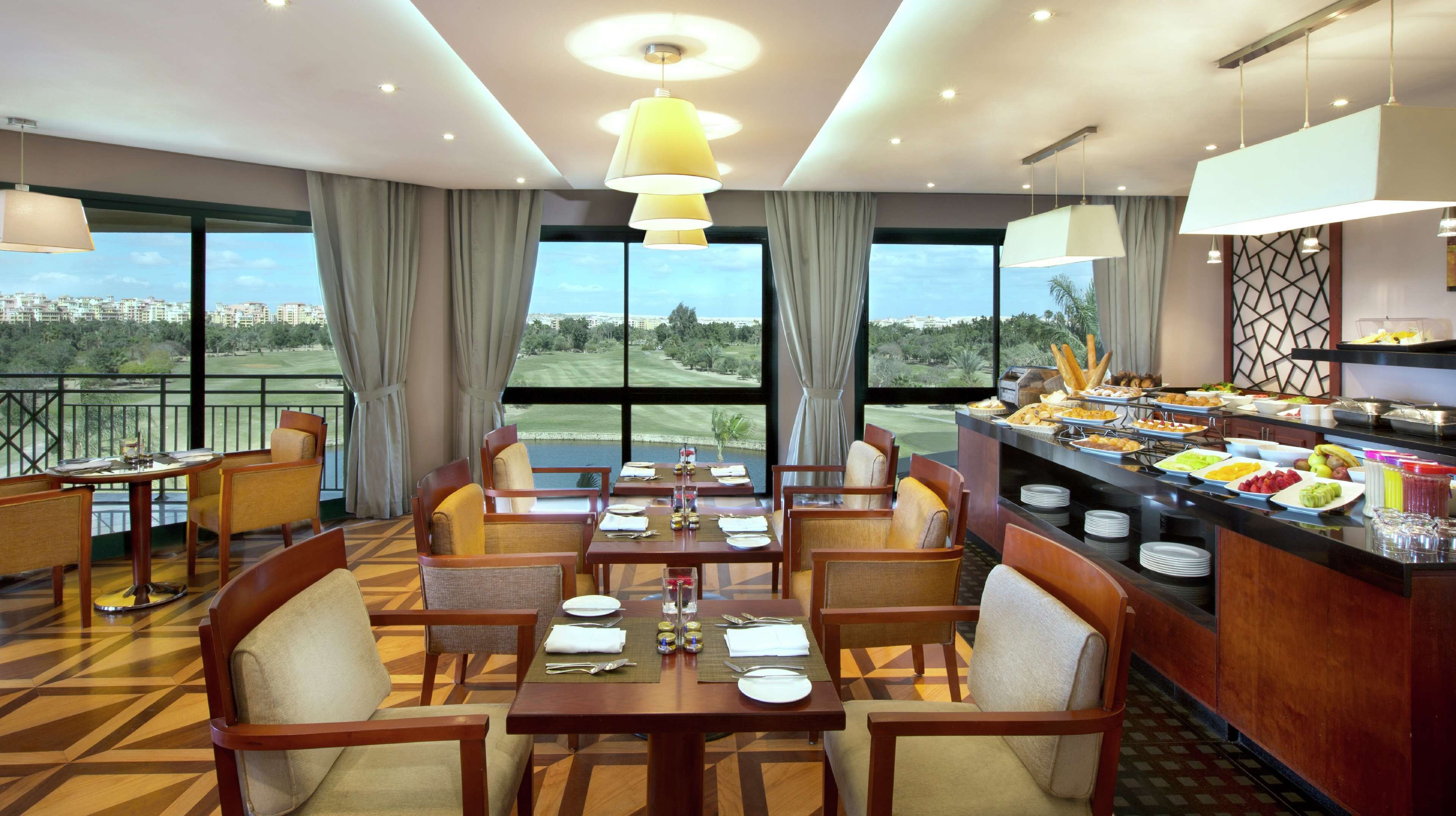 Une salle à manger d’hôtel avec des tables préparées pour le petit-déjeuner, de grandes fenêtres donnant sur un terrain de golf et un buffet proposant divers plats.