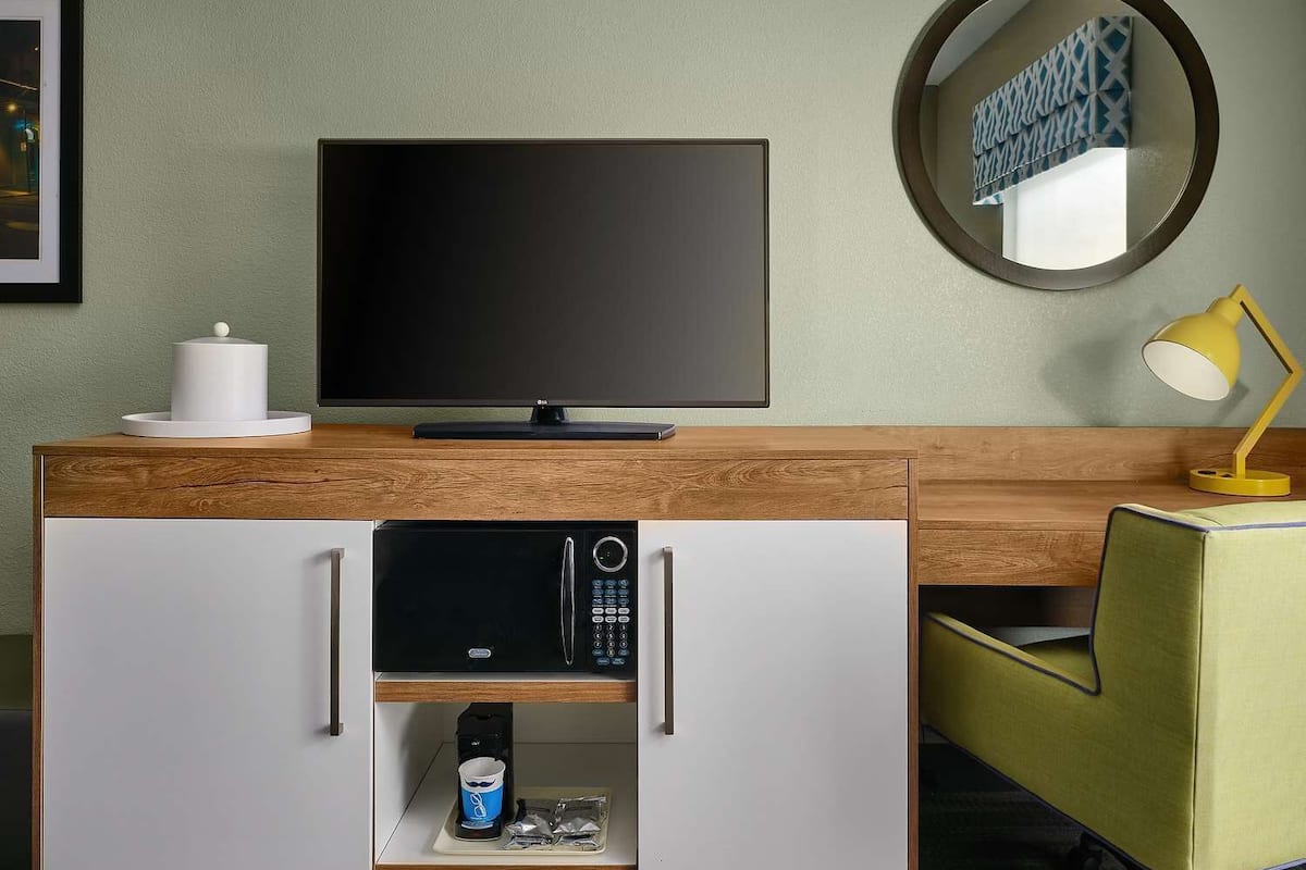A hotel room with a wooden desk, a flat-screen TV, a coffee machine, a microwave, a yellow desk lamp, and a green chair.