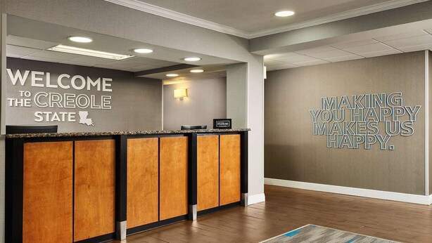 Een hotel receptie met een houten receptiebalie, een bord met de tekst 'Welcome to the Creole State', een muurspreuk met 'Making You Happy Makes Us Happy' en een vloerbedekking.