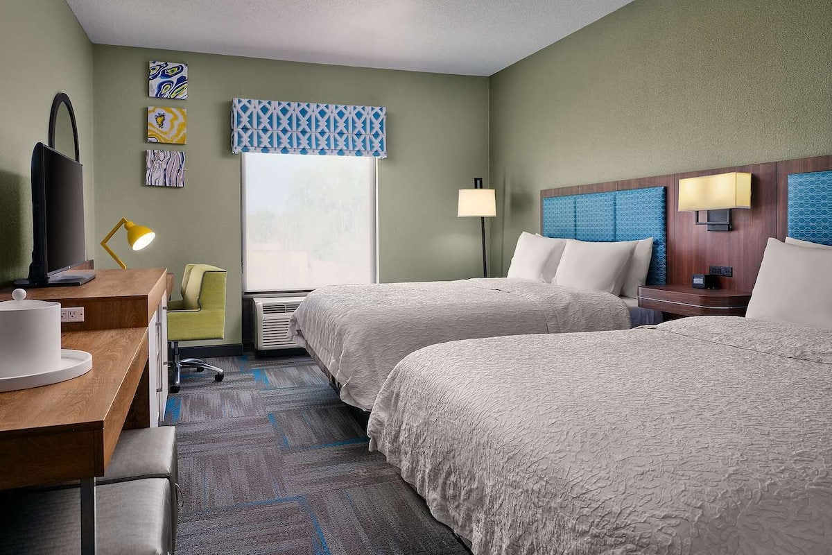A hotel room with two beds, a desk with a lamp, a chair, and artwork on the walls.