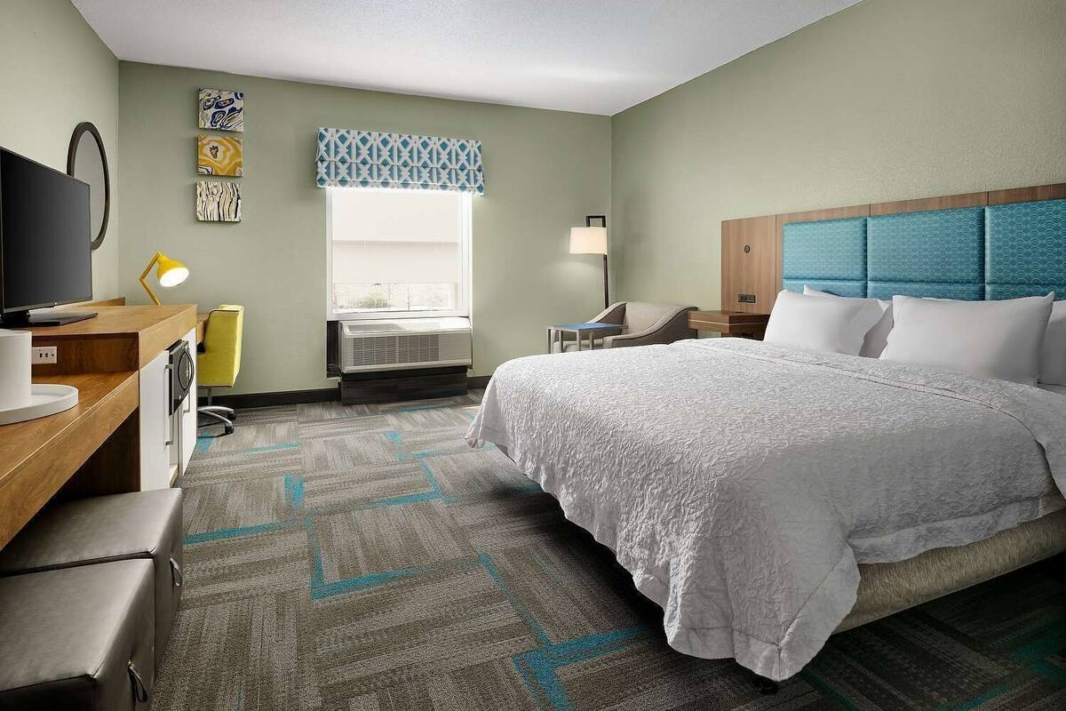 A hotel room with a bed, desk, chair, TV, lamp, and air conditioning unit.