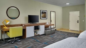 Een hotelkamer met een bed, een bureau met een stoel, een televisie, een spiegel, een lijst met een afbeelding en een deur.