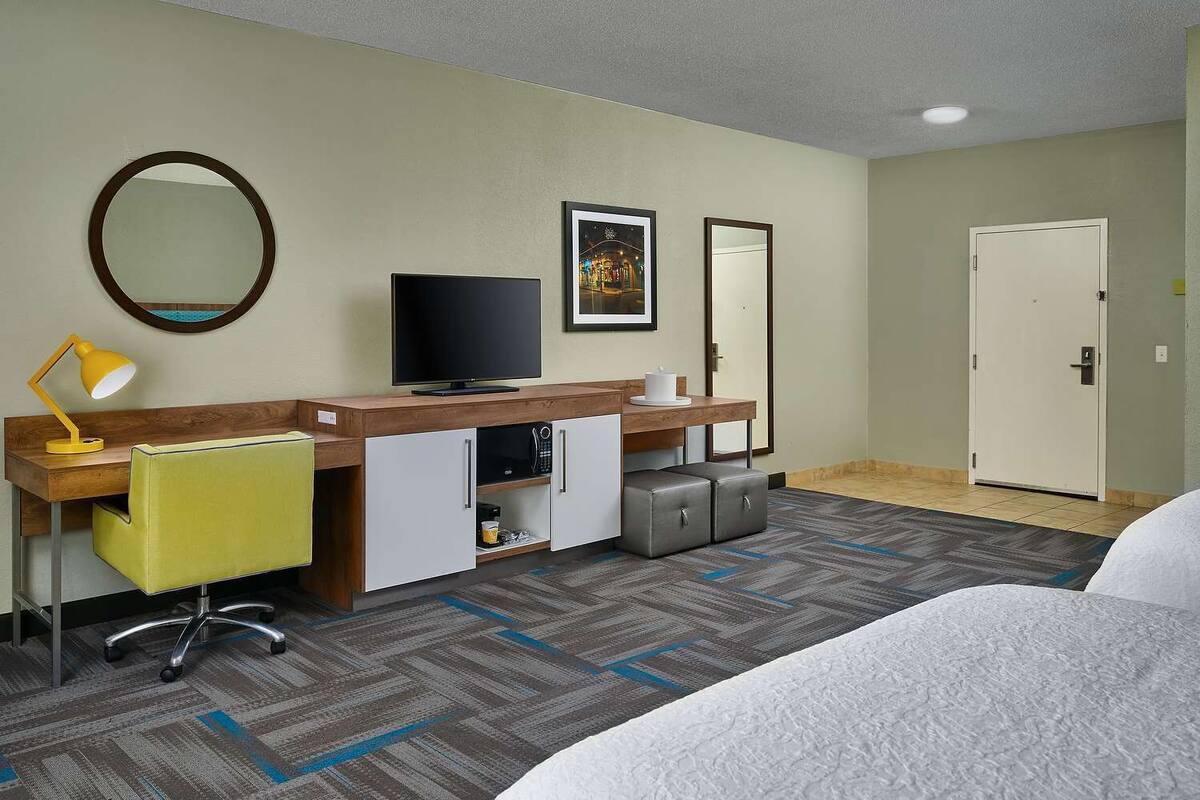 A hotel room with a bed, a desk with a chair, a TV, a mirror, a framed picture, and a door.