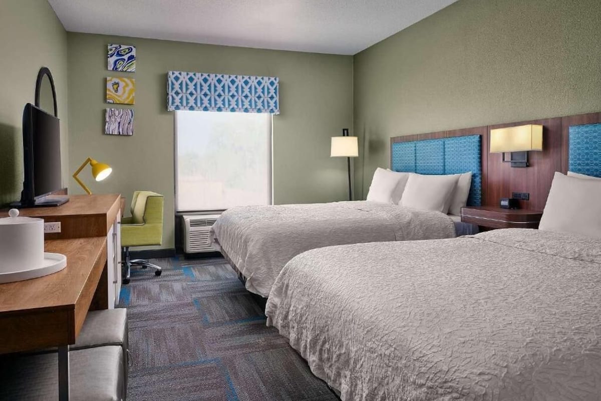 A hotel room with two beds, a desk with a lamp, a chair, and artwork on the walls.