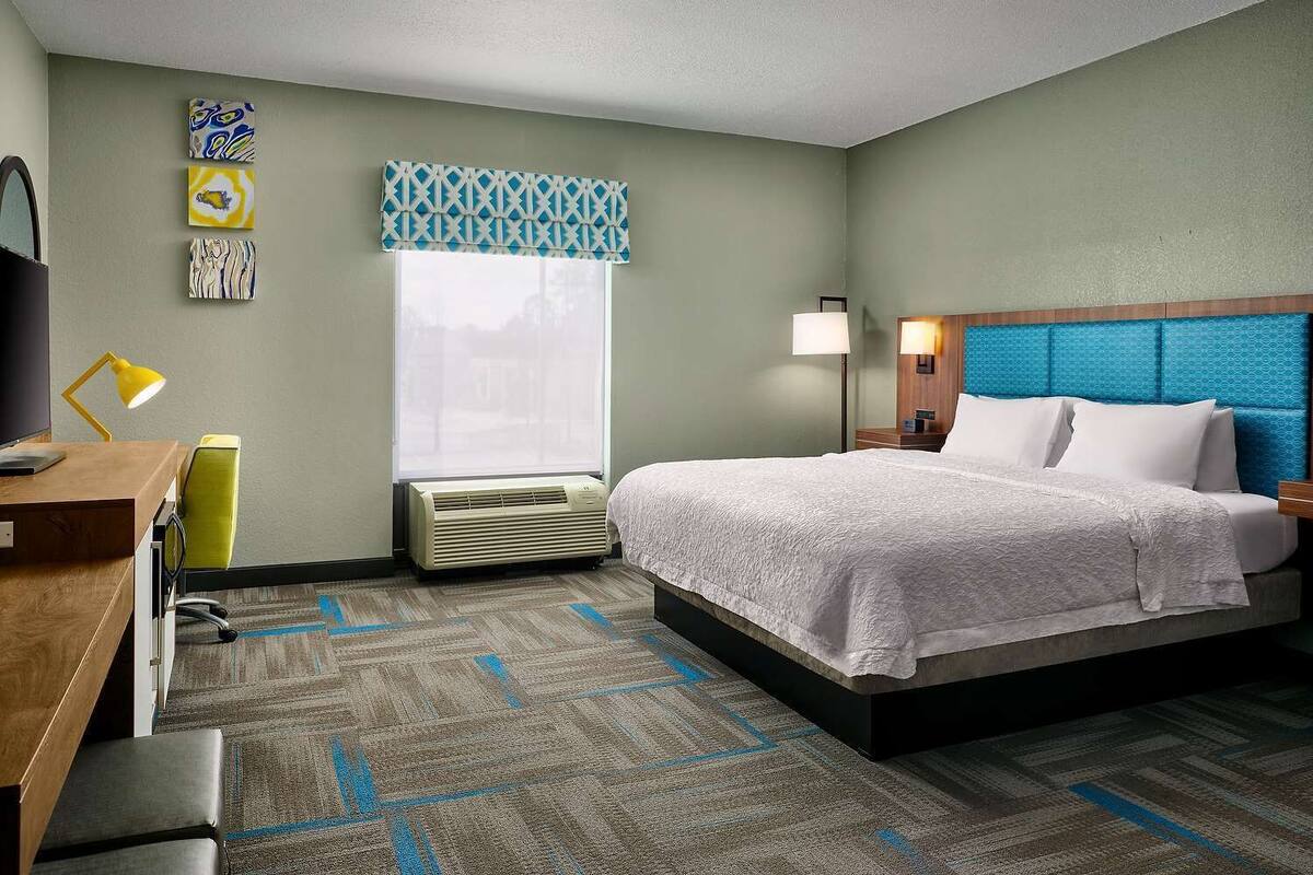 A hotel room with a bed, a desk with a chair, a TV, a lamp, and a window with a view.