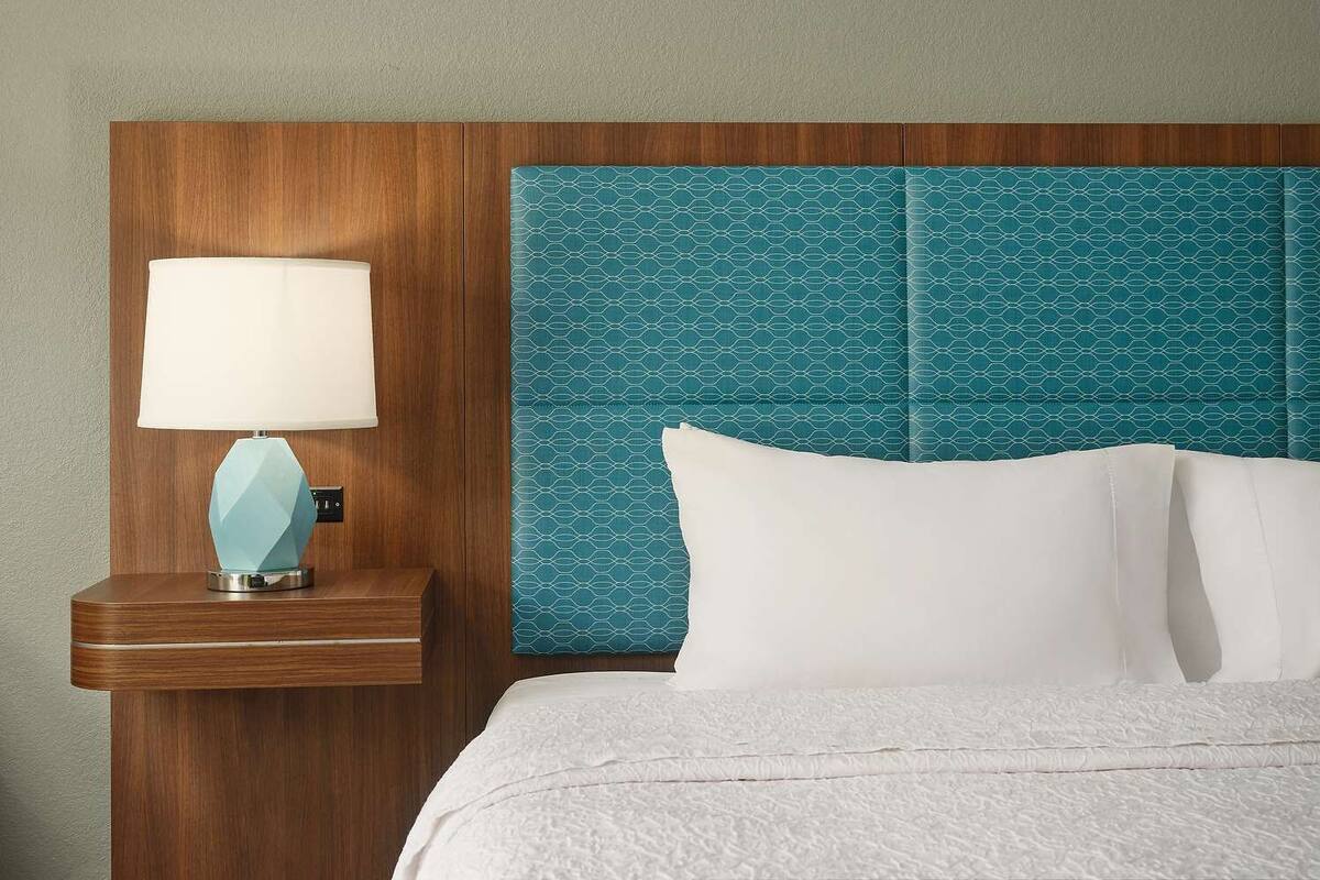 A hotel room with a wooden headboard, a nightstand with a lamp, and a bed with white bedding.