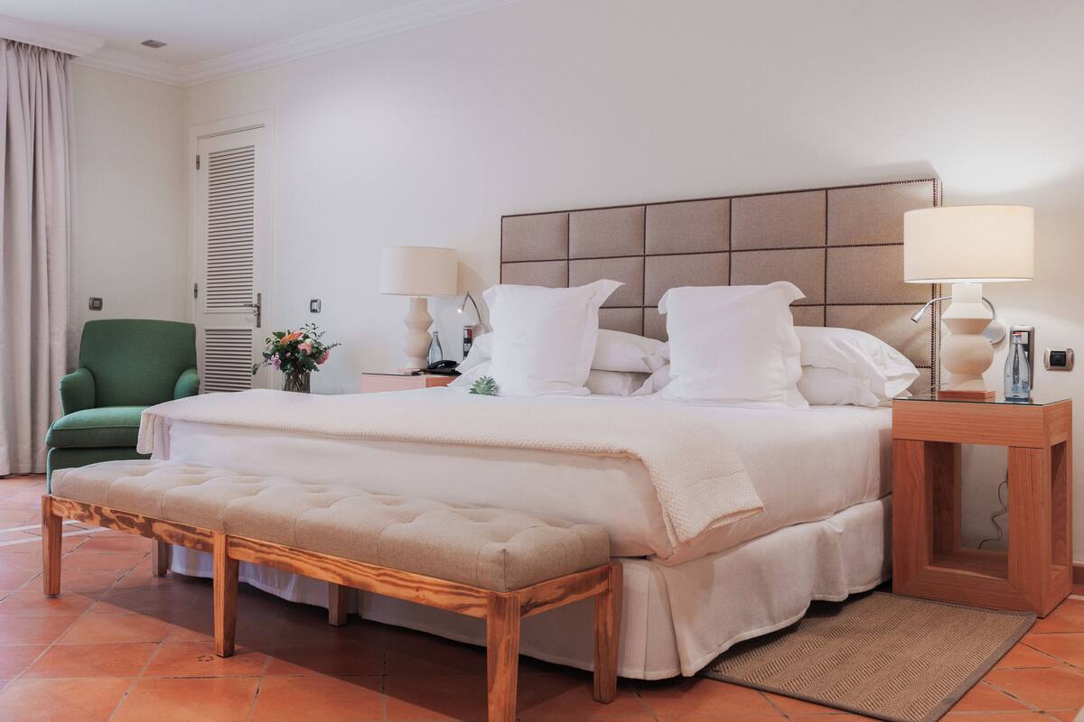A hotel room with a large bed, two bedside lamps, a green armchair, a wooden bench, and a tiled floor.