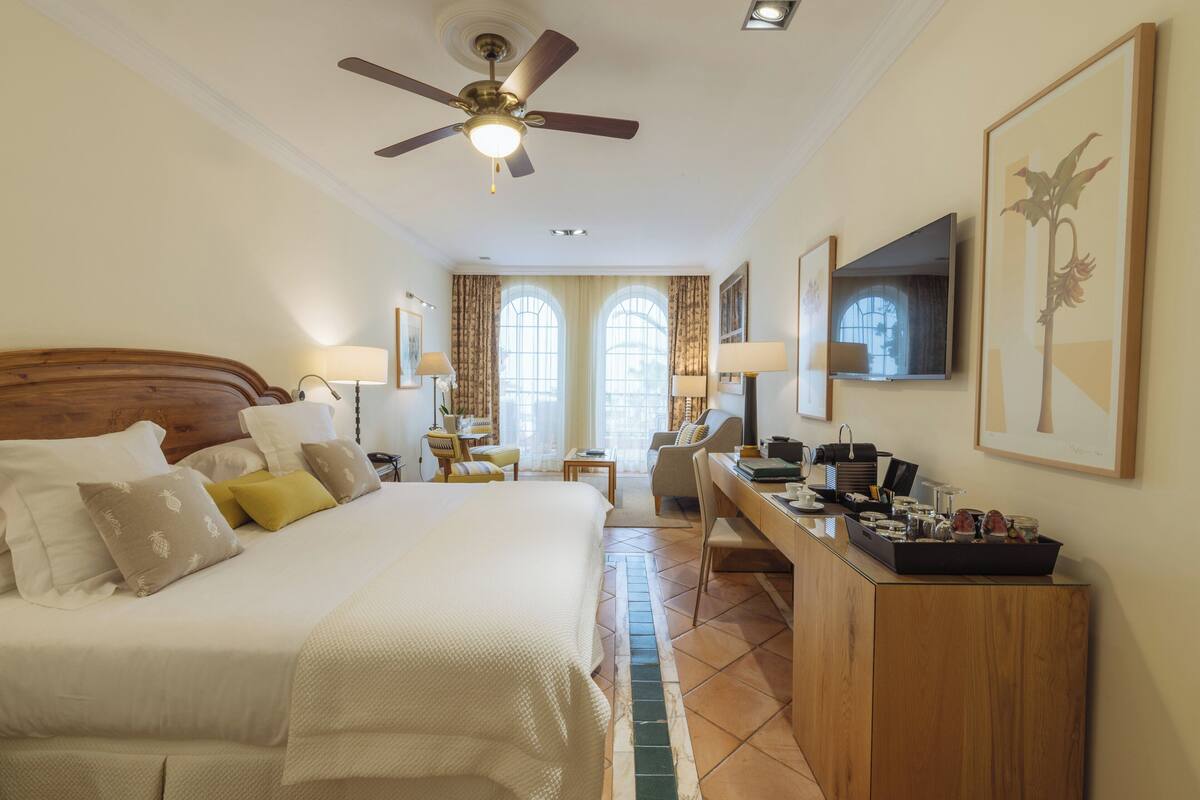 A hotel room with a large bed, a desk, a chair, a TV, and a ceiling fan.