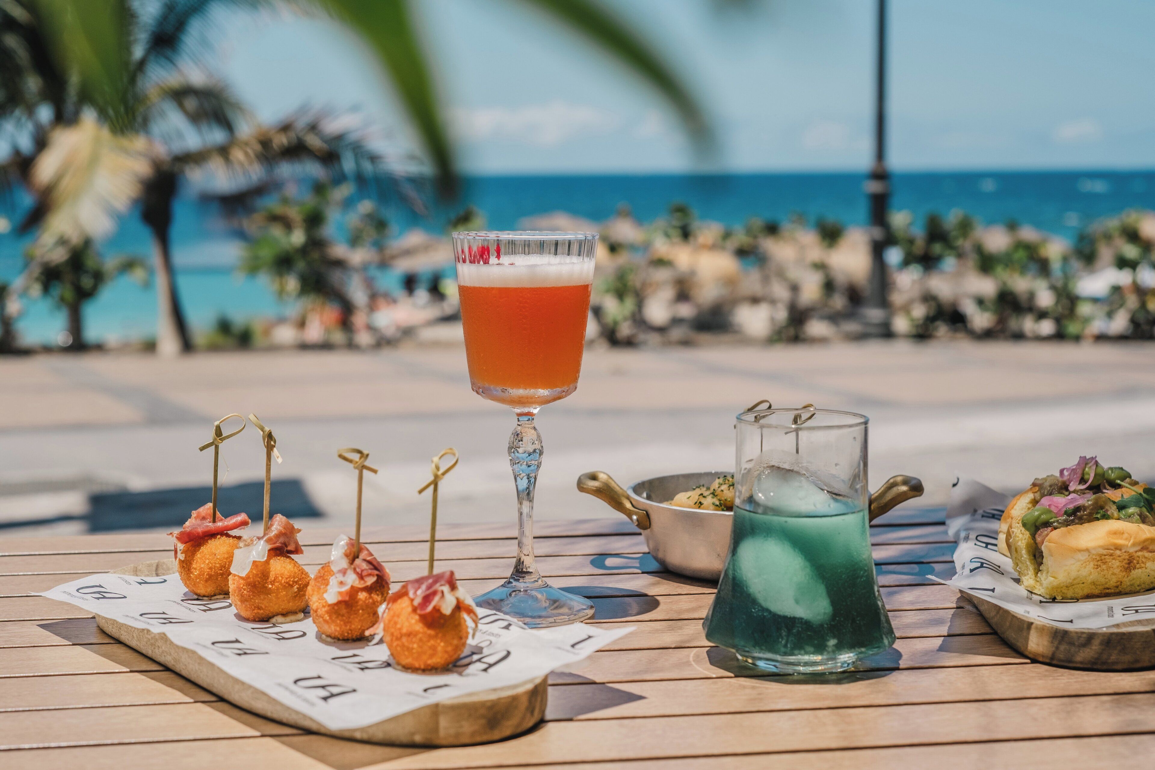 Una mesa con comida, una bebida y una jarrón sobre una terraza de madera con vistas al océano.