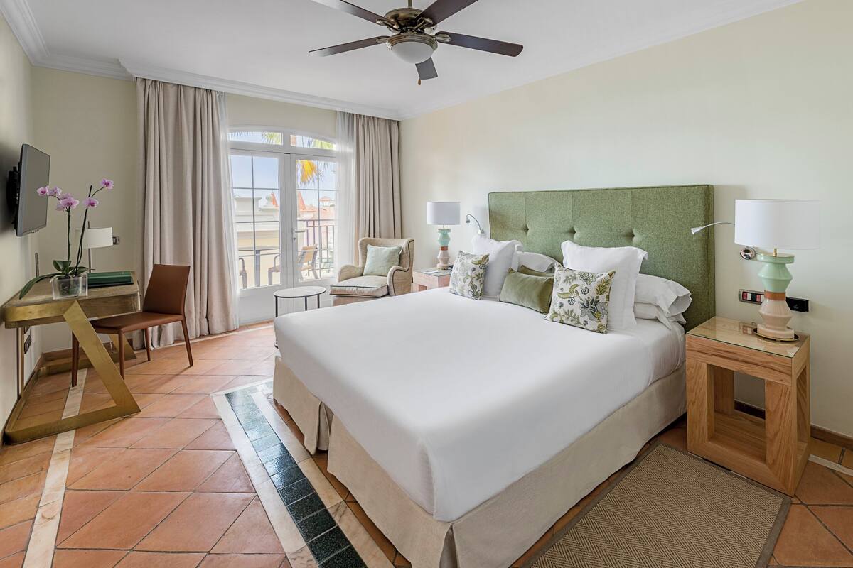 A hotel room with a large bed, a desk, a chair, a TV, and a balcony with curtains.