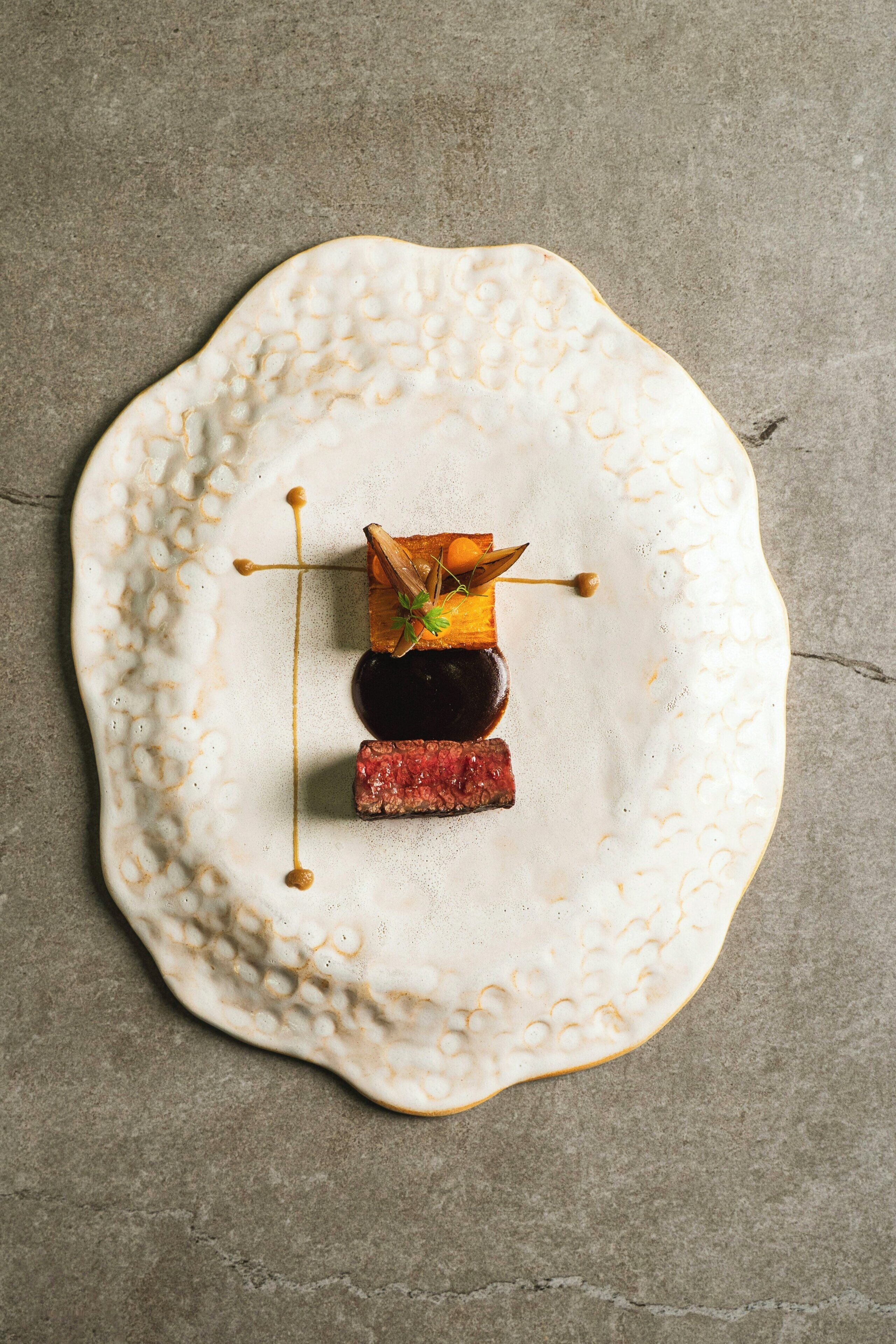 Un plato blanco y texturizado con un trozo de carne, una rodaja de berenjena y un adorno sobre una superficie gris.
