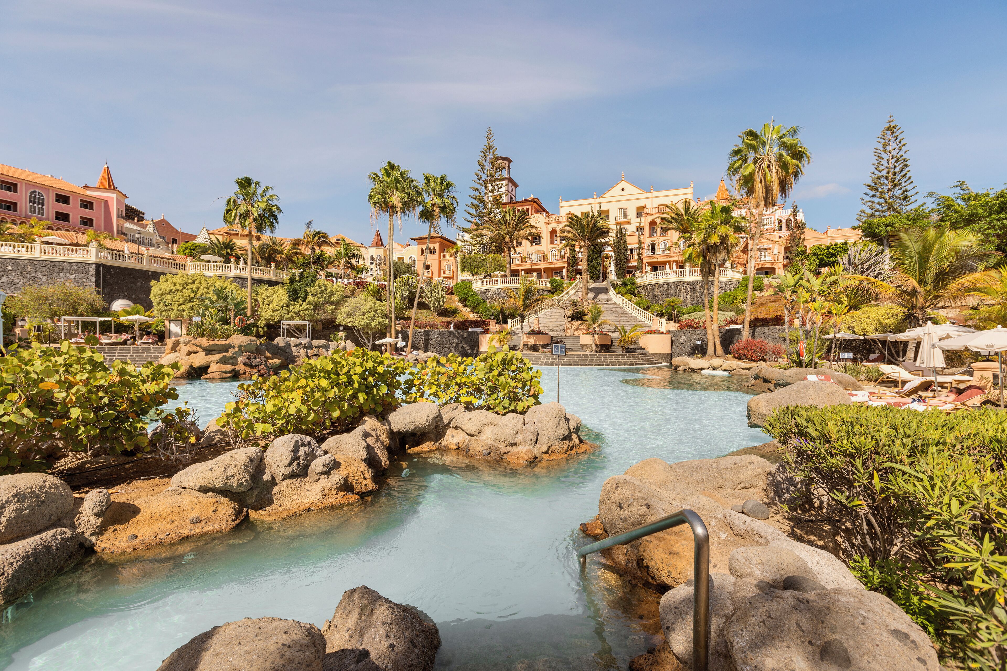 Un complejo turístico con piscina rodeada de rocas y palmeras, y un edificio grande al fondo.