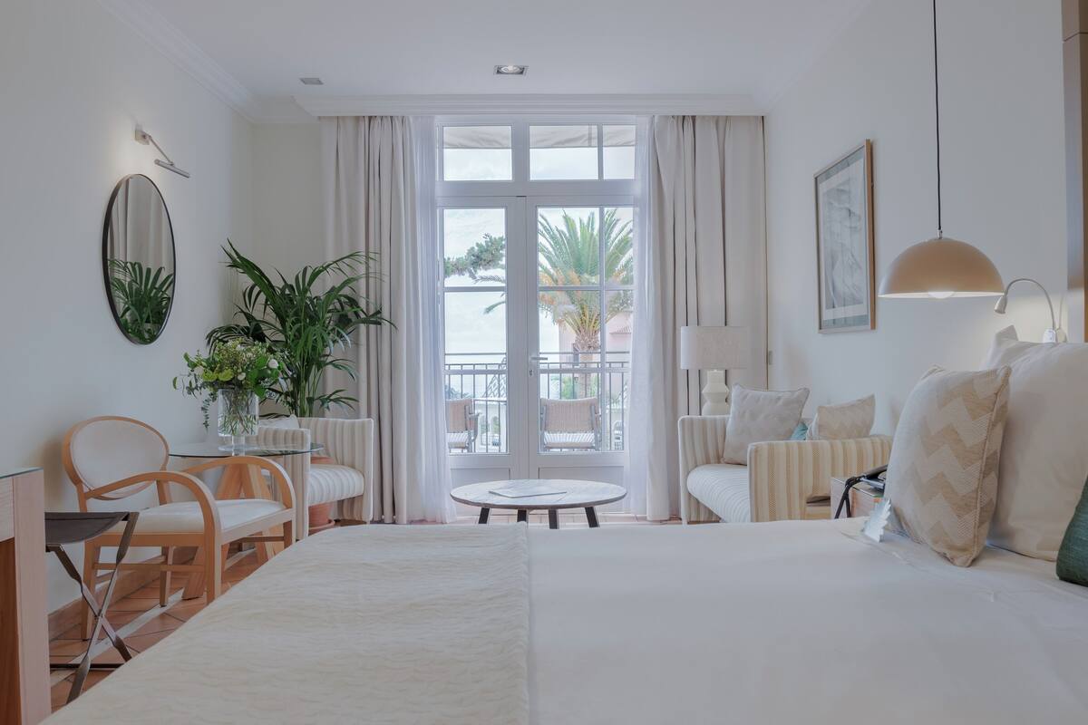 A hotel room with a large bed, two armchairs, a small table, a potted plant, and a view of the outside balcony.
