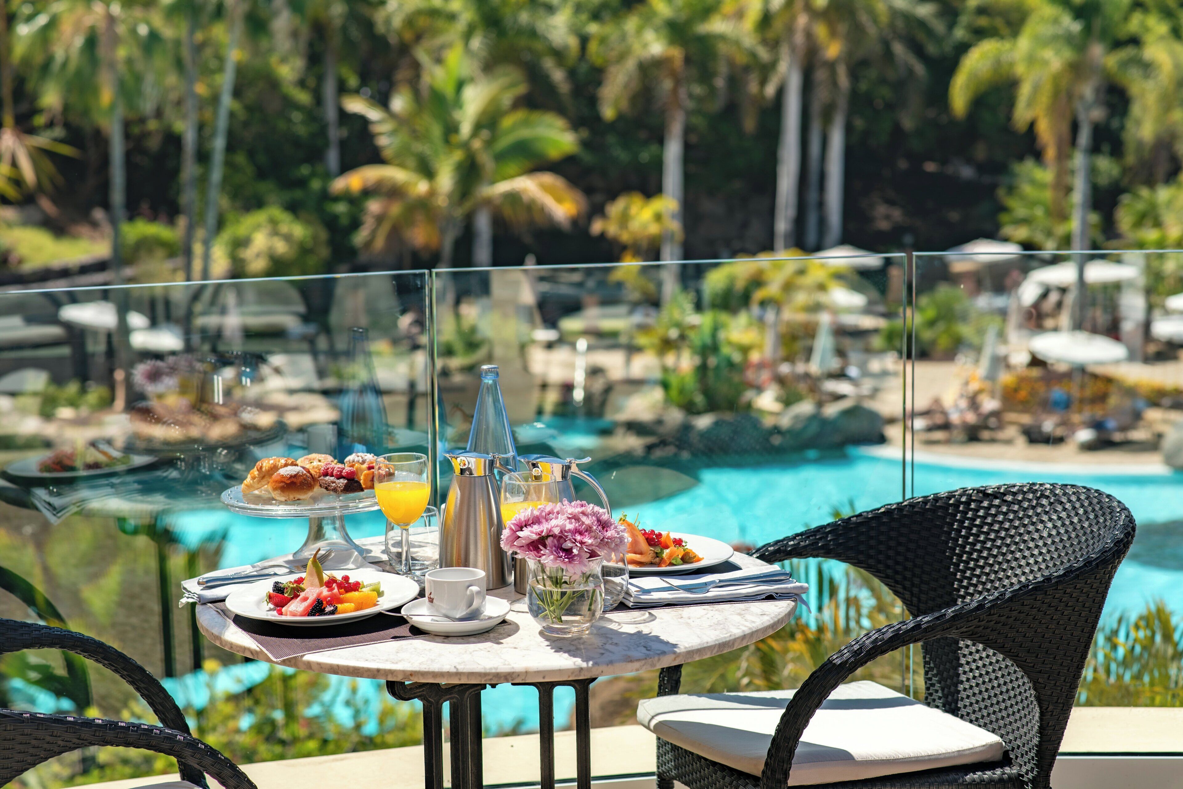 Ein Esstisch mit Frühstücksangeboten, einer Blumenvase und Blick auf ein Schwimmbad und Palmen.
