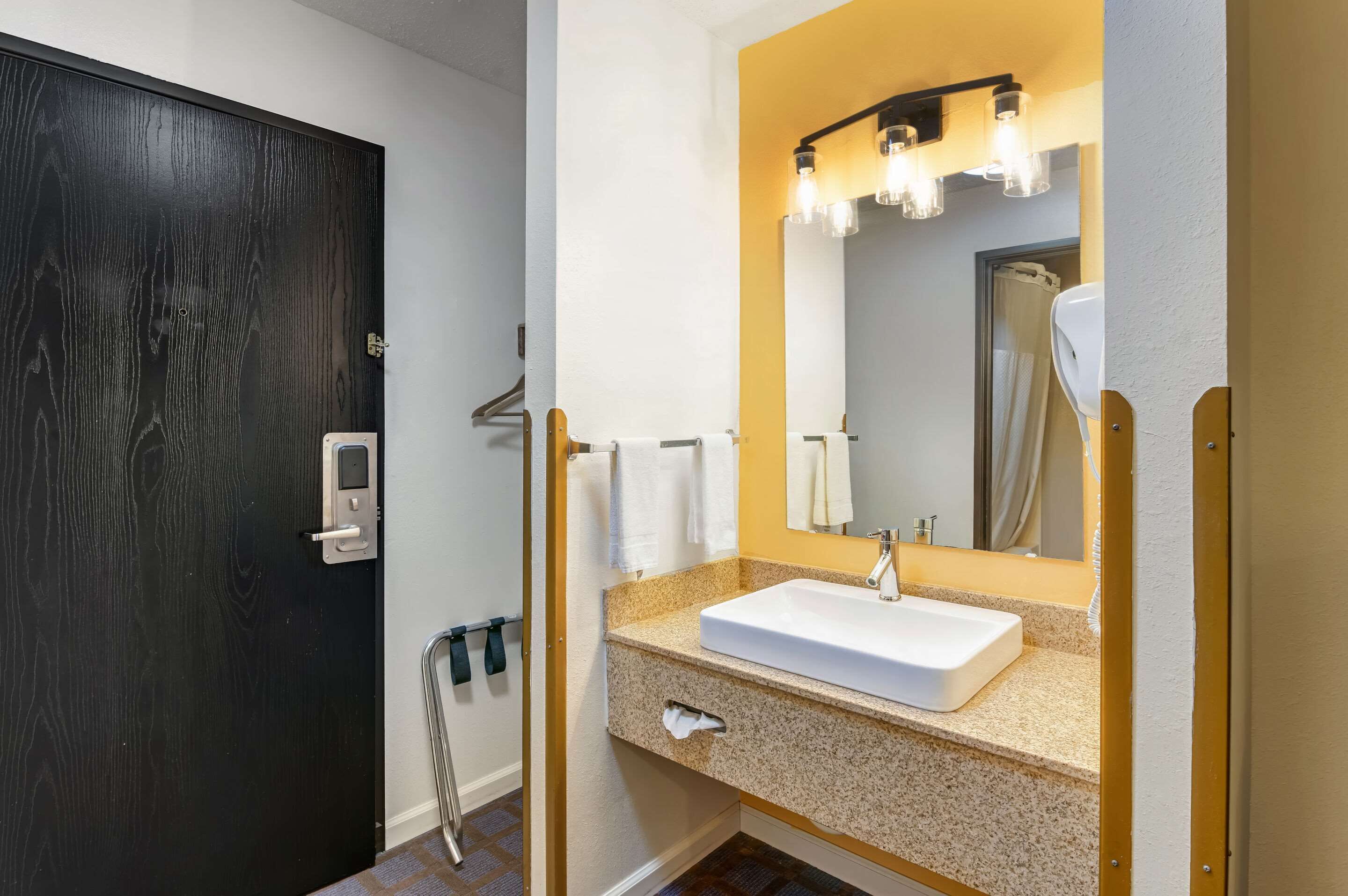 A bathroom with a dark wooden door, a white sink with a beige countertop, a mirror with lights, and a towel rack.