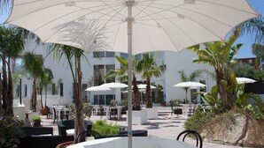 Un área de patio al aire libre con mesas y sillas blancas, rodeada de palmeras y plantas en macetas.