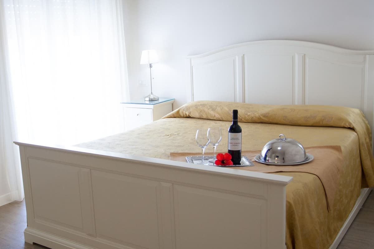 A neatly made bed with a white headboard, a bedside table with a lamp, and a bottle of wine on the bed.