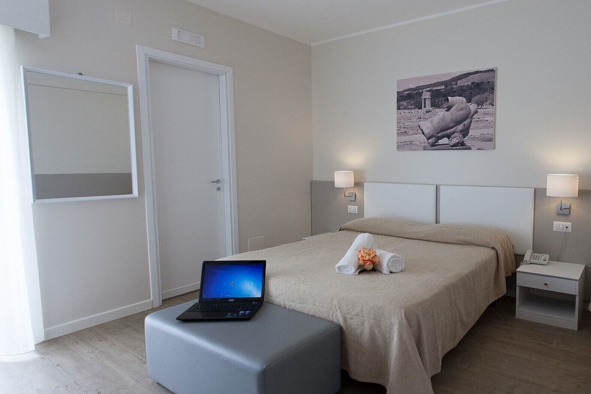 A hotel room with a bed, a desk with a laptop, a bedside table with a lamp, and a wall-mounted picture.