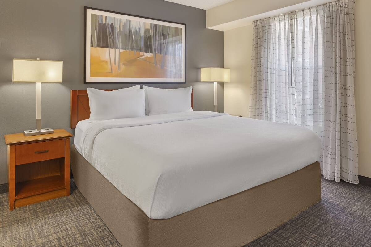 A neatly made bed with white linens, a wooden nightstand with a lamp, a wall-mounted painting, and a window with curtains.
