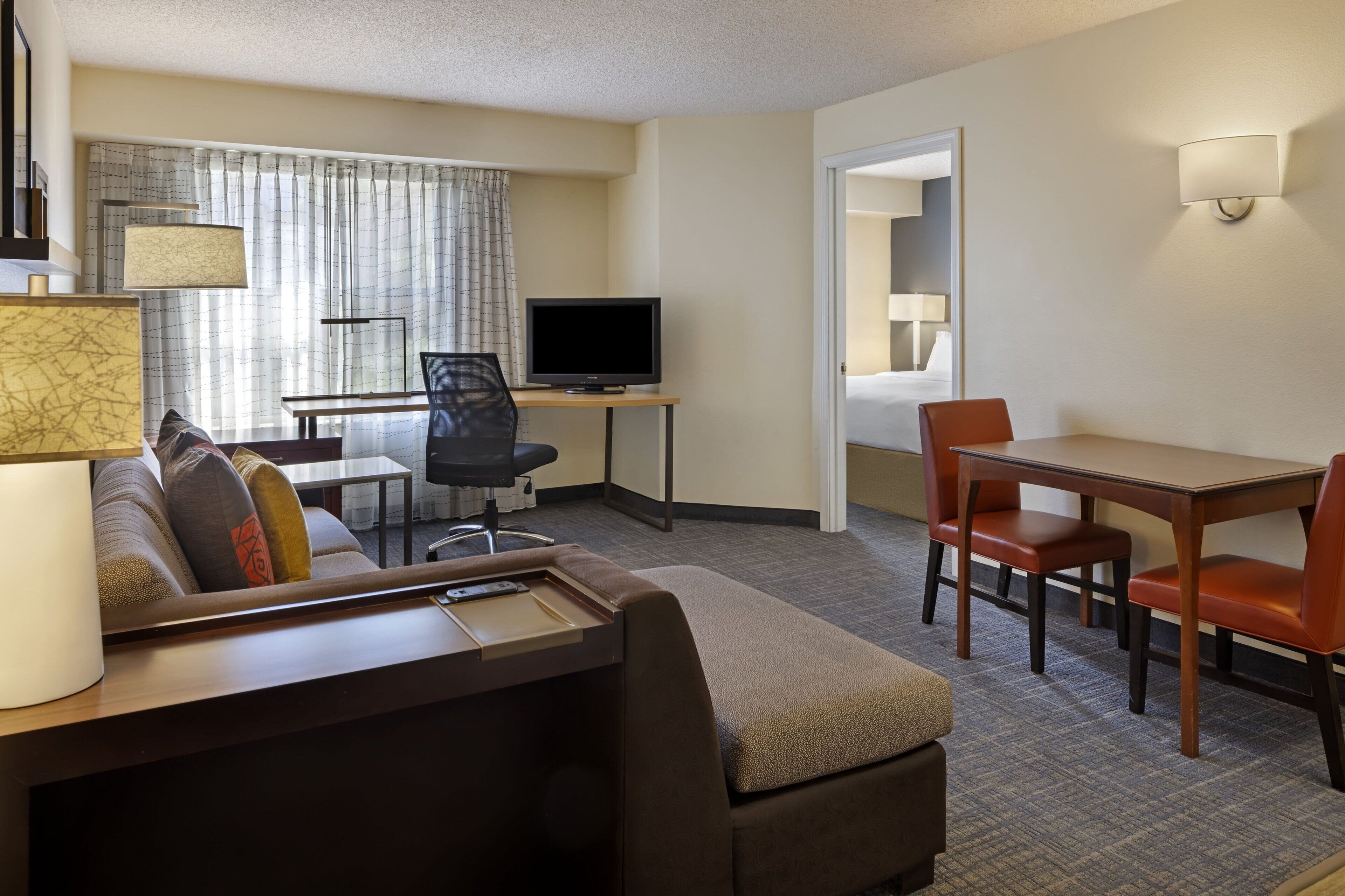A hotel room with a sofa, a small table with a lamp, a TV, and a dining area with chairs.