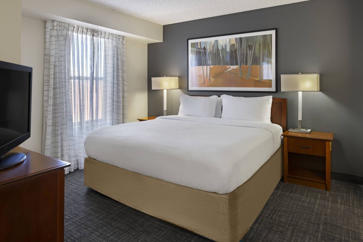 A hotel room with a large bed, two bedside tables with lamps, a flat-screen TV, and a painting on the wall.