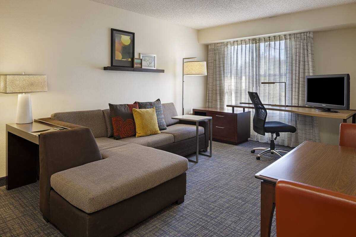A hotel room with a sofa, ottoman, desk, chair, television, lamp, and a window with curtains.