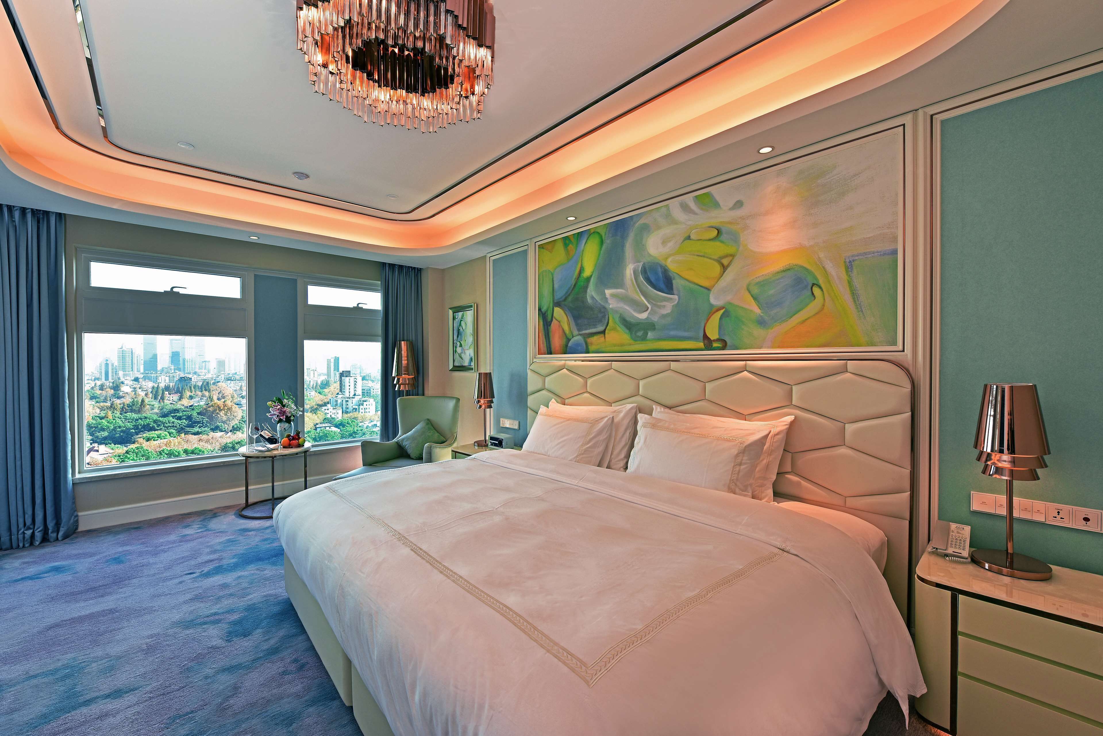 A modern hotel room with a large bed, a city view, a unique abstract painting, and a chandelier.