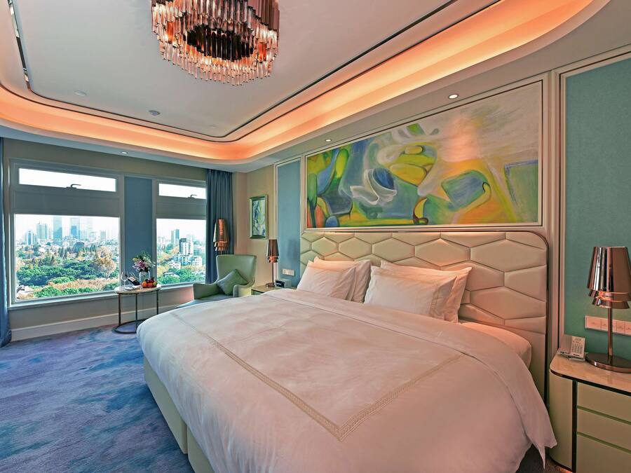 A modern hotel room with a large bed, a city view, a unique abstract painting, and a chandelier.