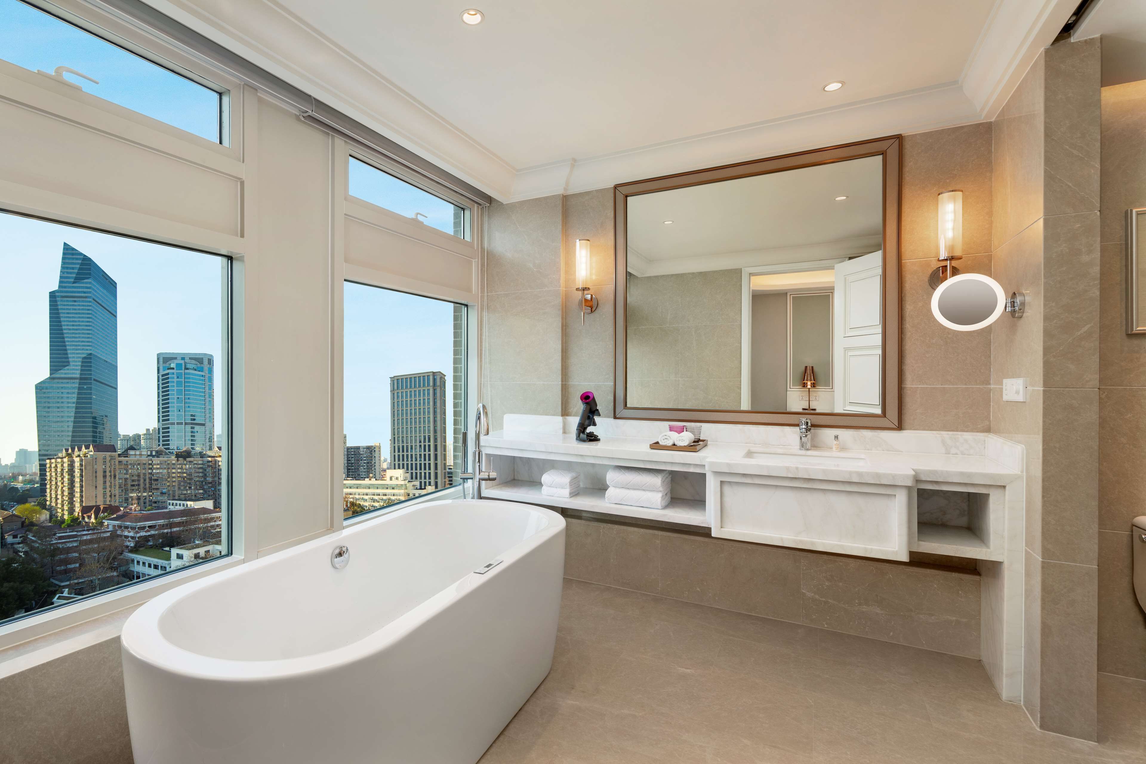 A modern bathroom with a large bathtub, a vanity with a mirror, and a view of the cityscape through large windows.
