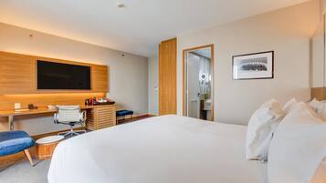 Quarto de hotel com uma cama grande, uma escrivaninha com cadeira, uma televisão na parede e um quadro emoldurado.