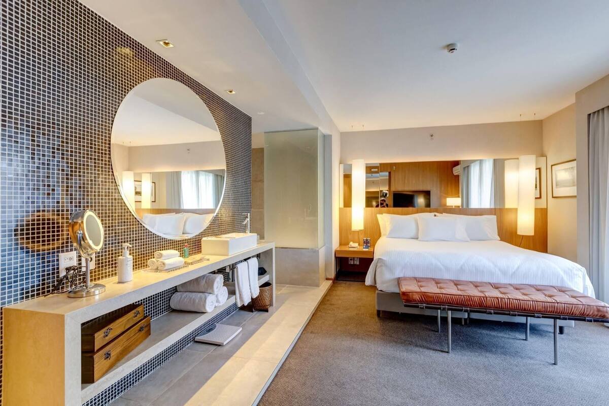 A modern hotel room with a large mirror, a bed, a bench, and a vanity with toiletries.