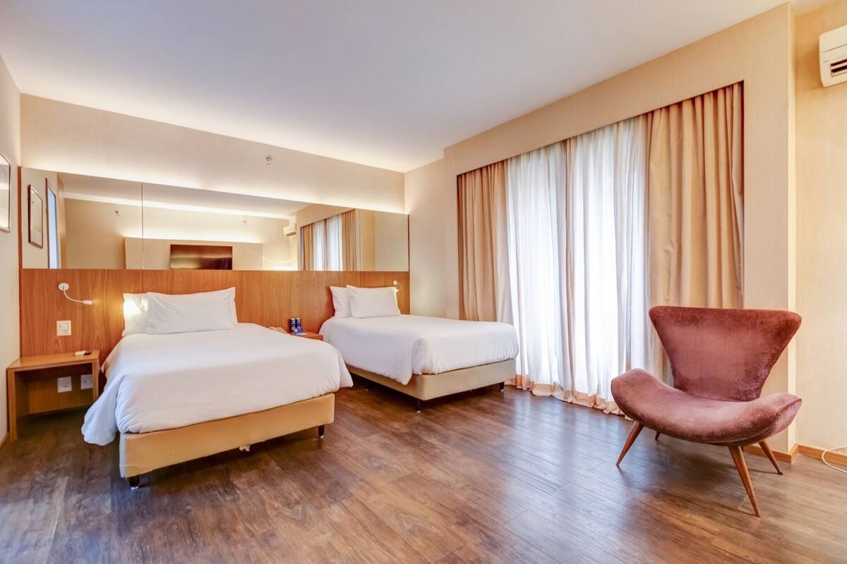 A hotel room with two beds, a wooden headboard, a large mirror, a chair, and a window with curtains.