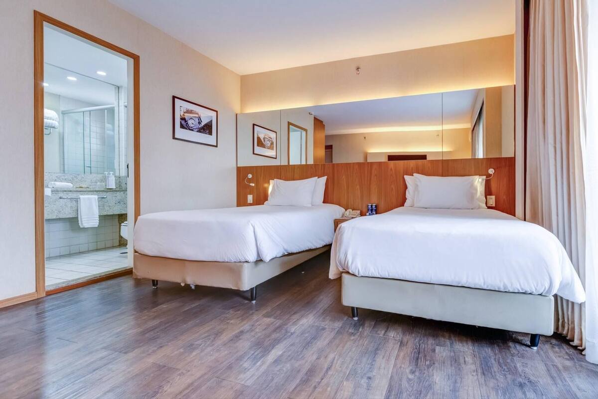 A hotel room with two beds, a bathroom with a glass shower, and a wooden headboard.