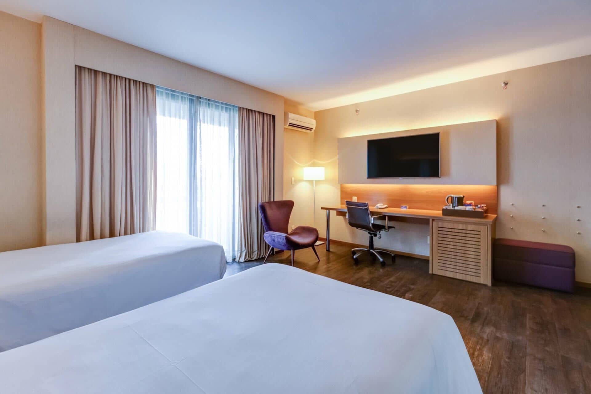 A hotel room with two beds, a desk with a chair, a TV, and a lamp.