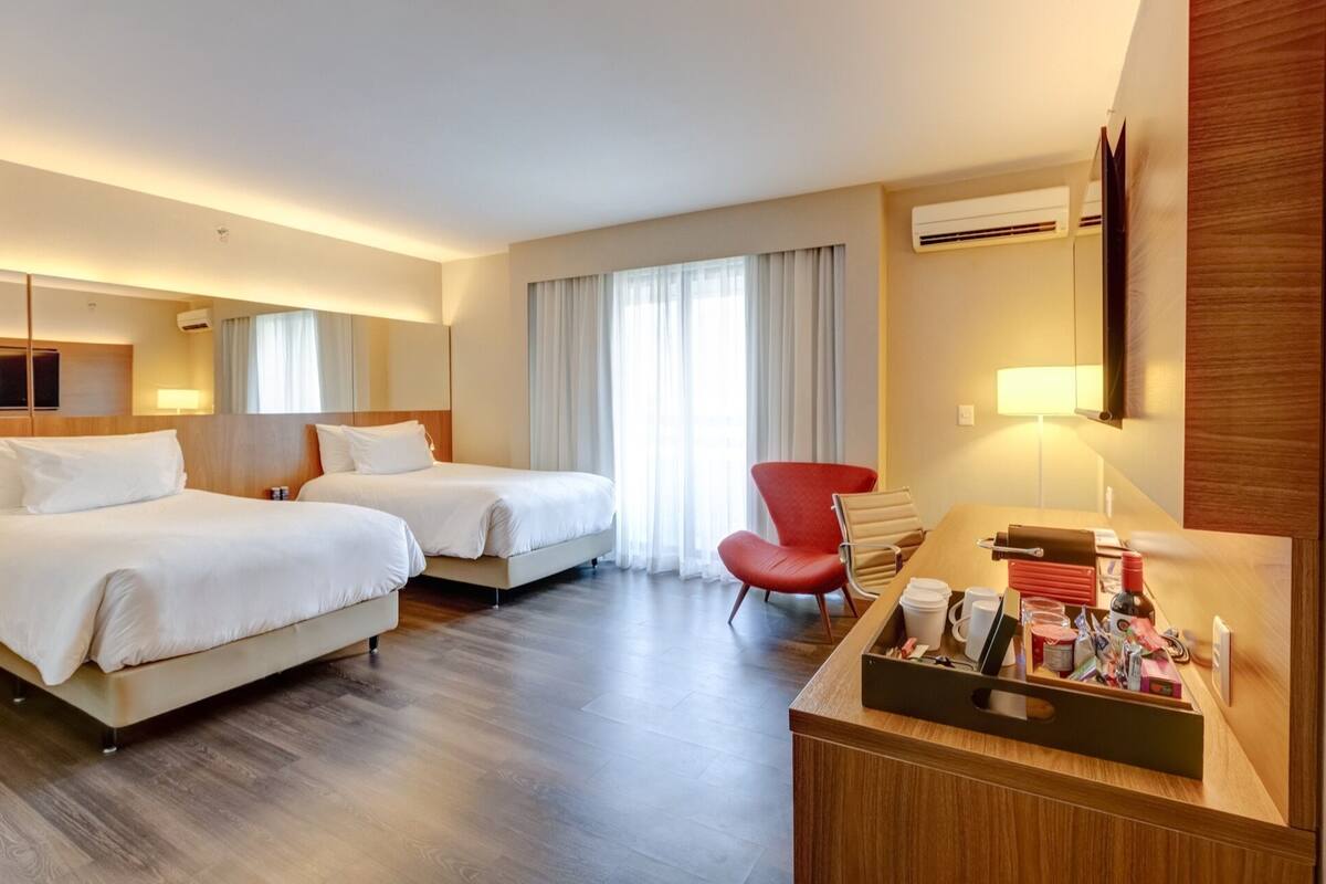 A hotel room with two beds, a desk with amenities, a red chair, and a large window with curtains.