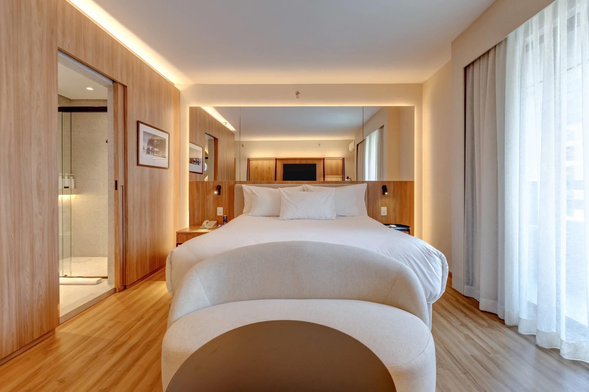Een moderne hotelkamer met een groot bed, houten vloeren, een flatscreen televisie en een groot raam met gordijnen.