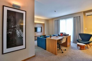 Ein Hotelzimmer mit Stadtbild-Kunstwerk, einem Holztisch, einem blauen Sofa, einem Fernseher und einem großen Fenster mit Vorhängen.