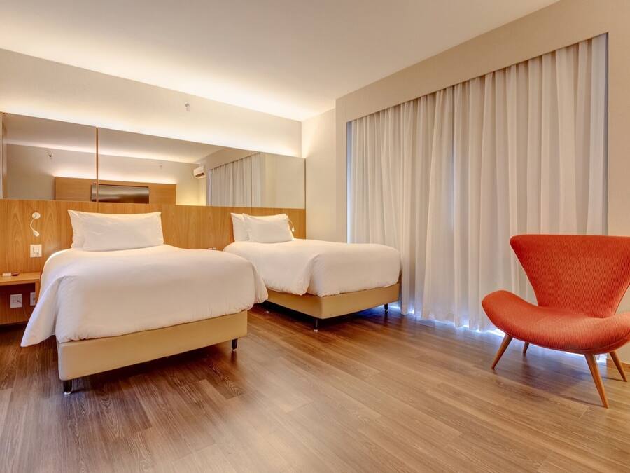 A hotel room with two beds, a red chair, wooden flooring, and a large window with curtains.