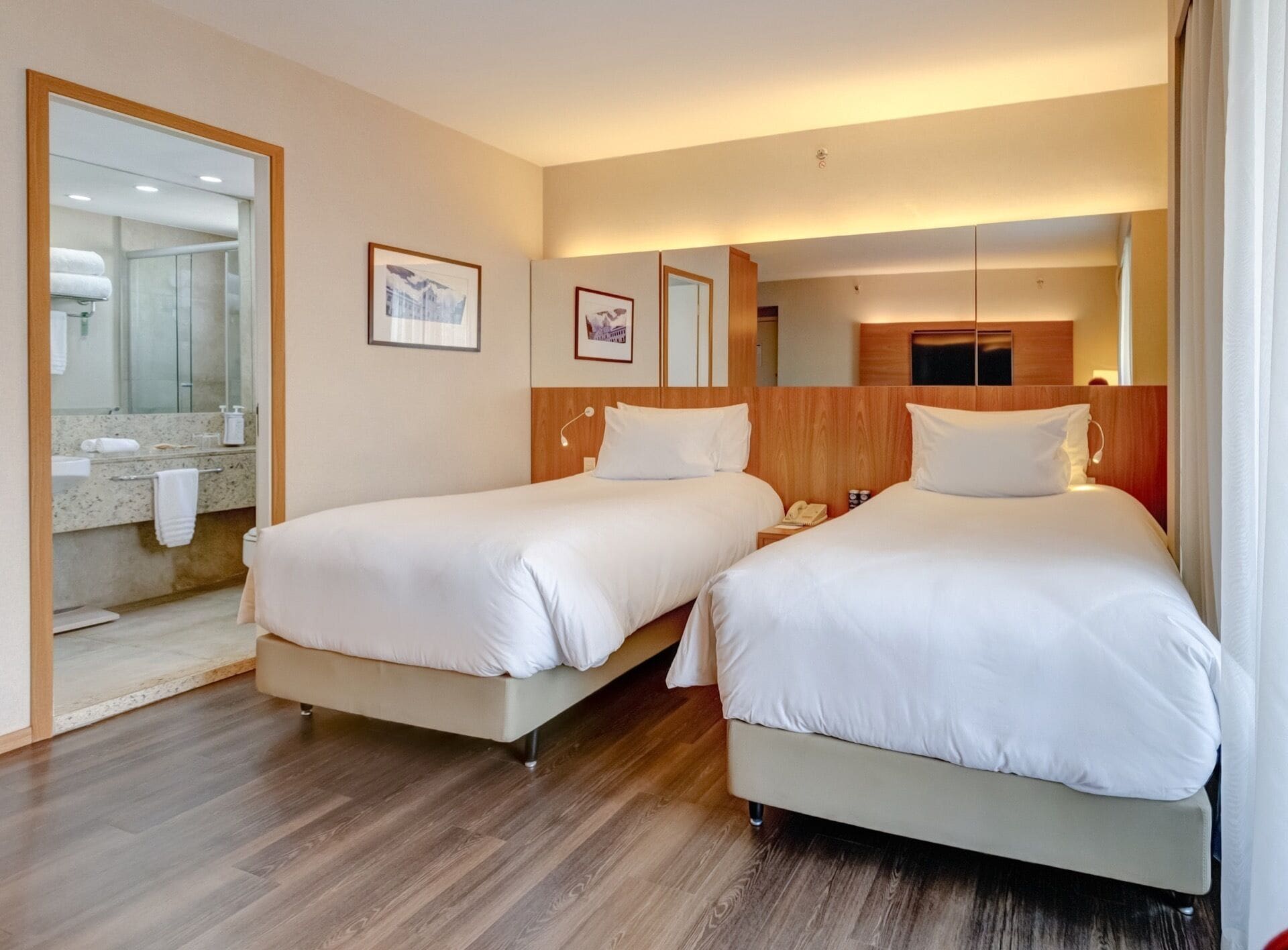 A hotel room with two beds, a bathroom with a mirror, and a wooden floor.