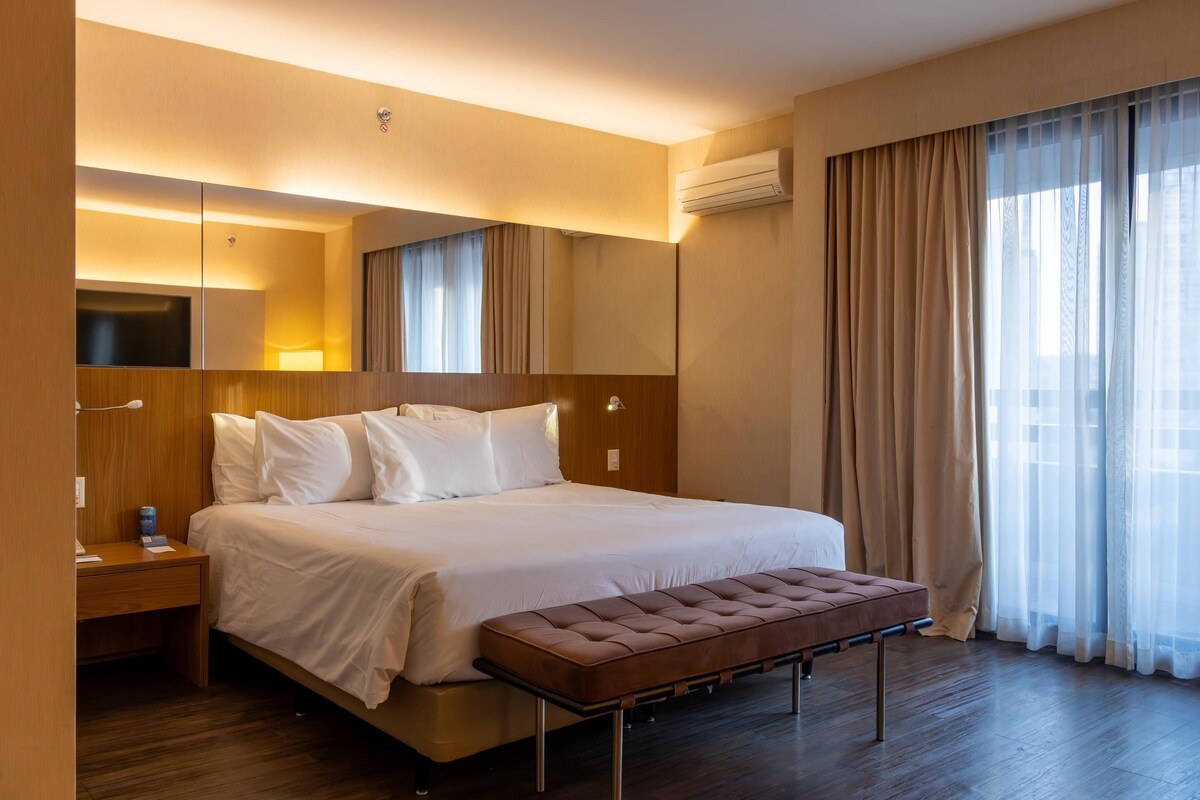 A hotel room with a large bed, a bedside table, a bench, a mirror, and a window with curtains.