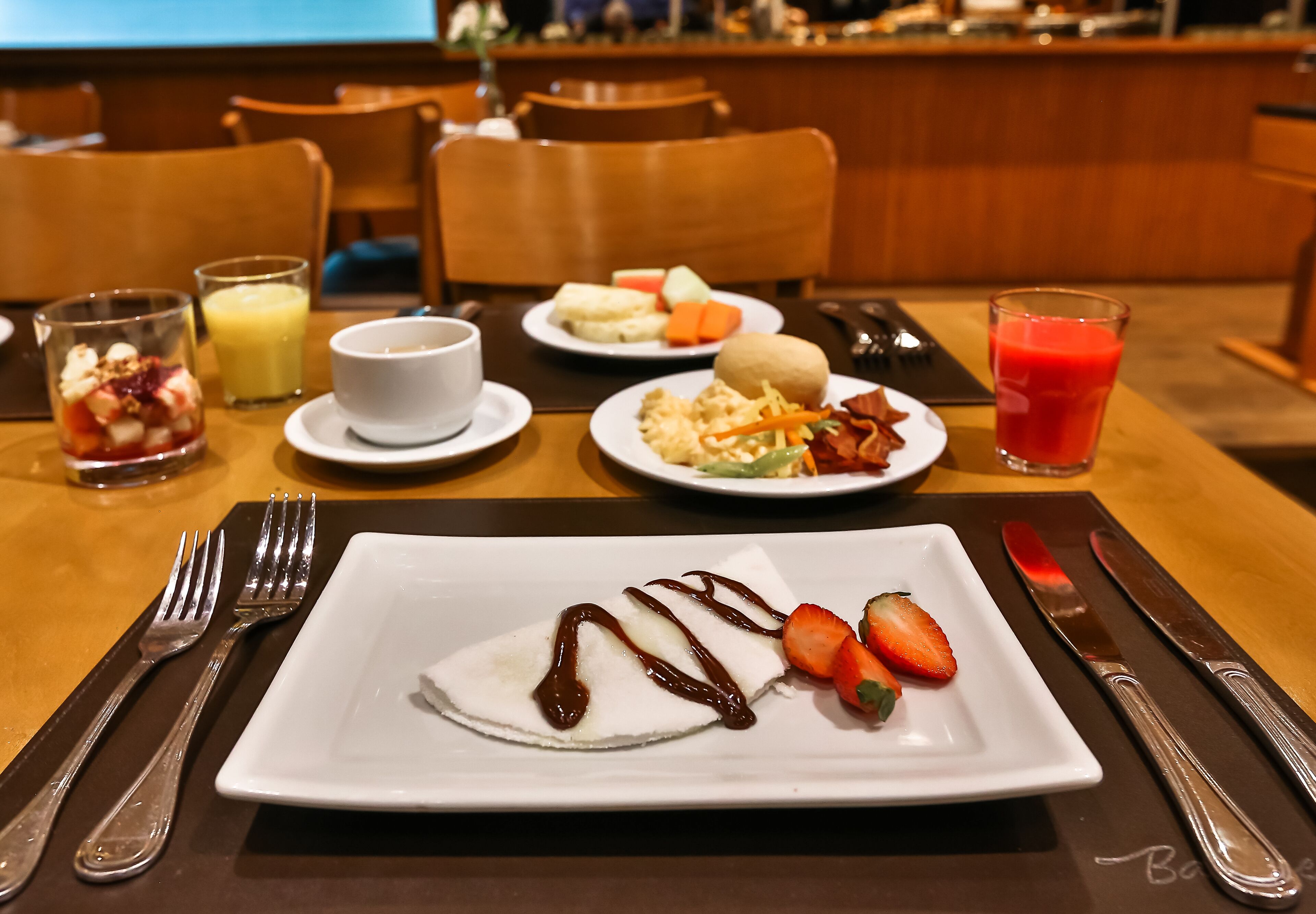 A breakfast setting with a plate of pancakes, strawberries, and chocolate sauce, a glass of orange juice, and a plate of eggs and bacon.