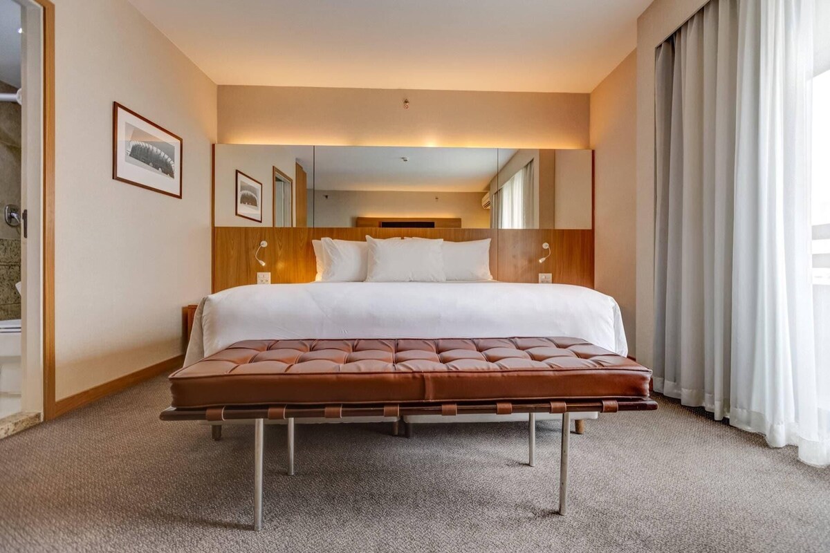 A hotel room with a large bed, a wooden headboard, a tufted bench, and a large window with curtains.