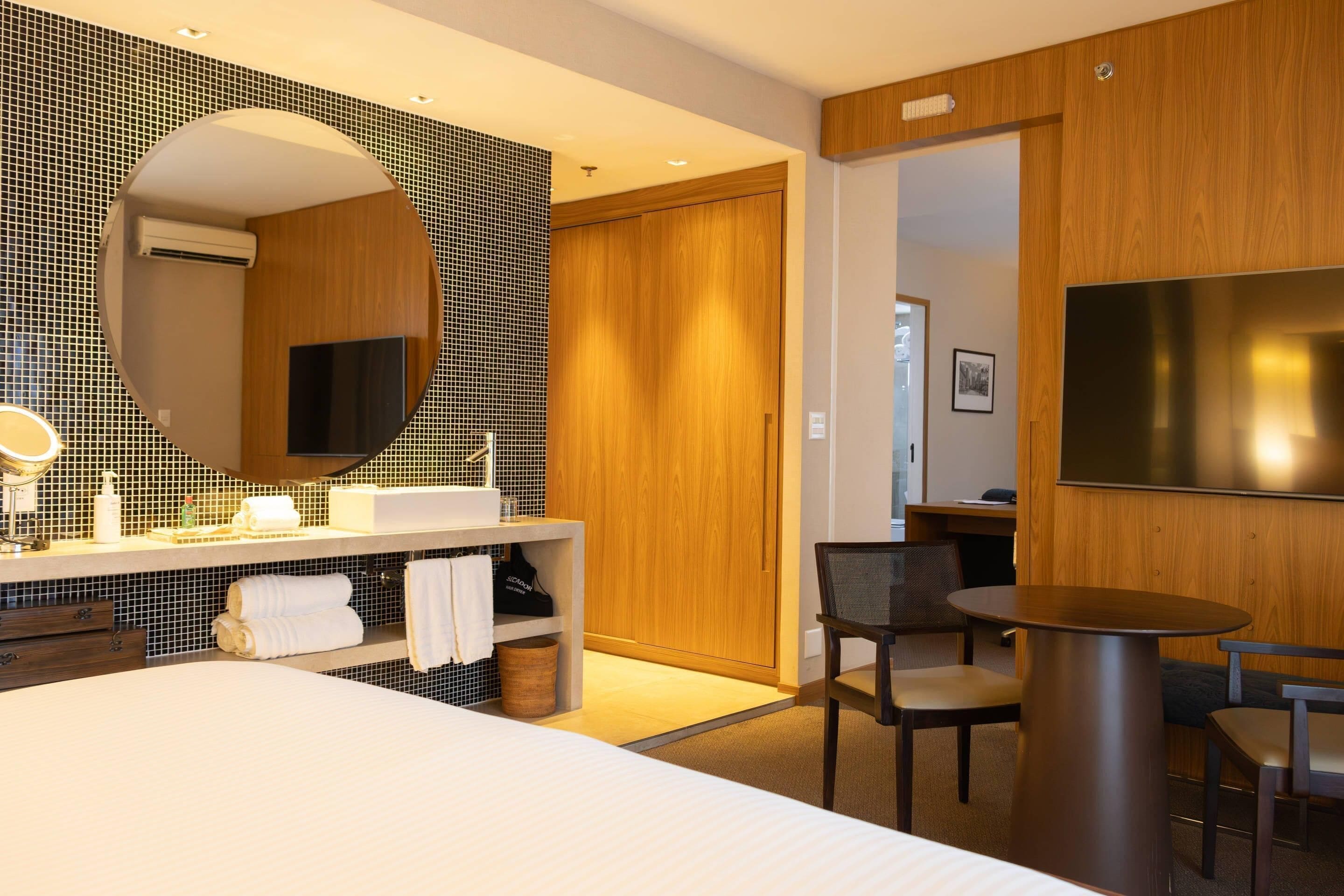 A modern hotel room with a large bed, a round dining table, a wooden wardrobe, a TV, and a bathroom with a tiled backsplash and a large mirror.
