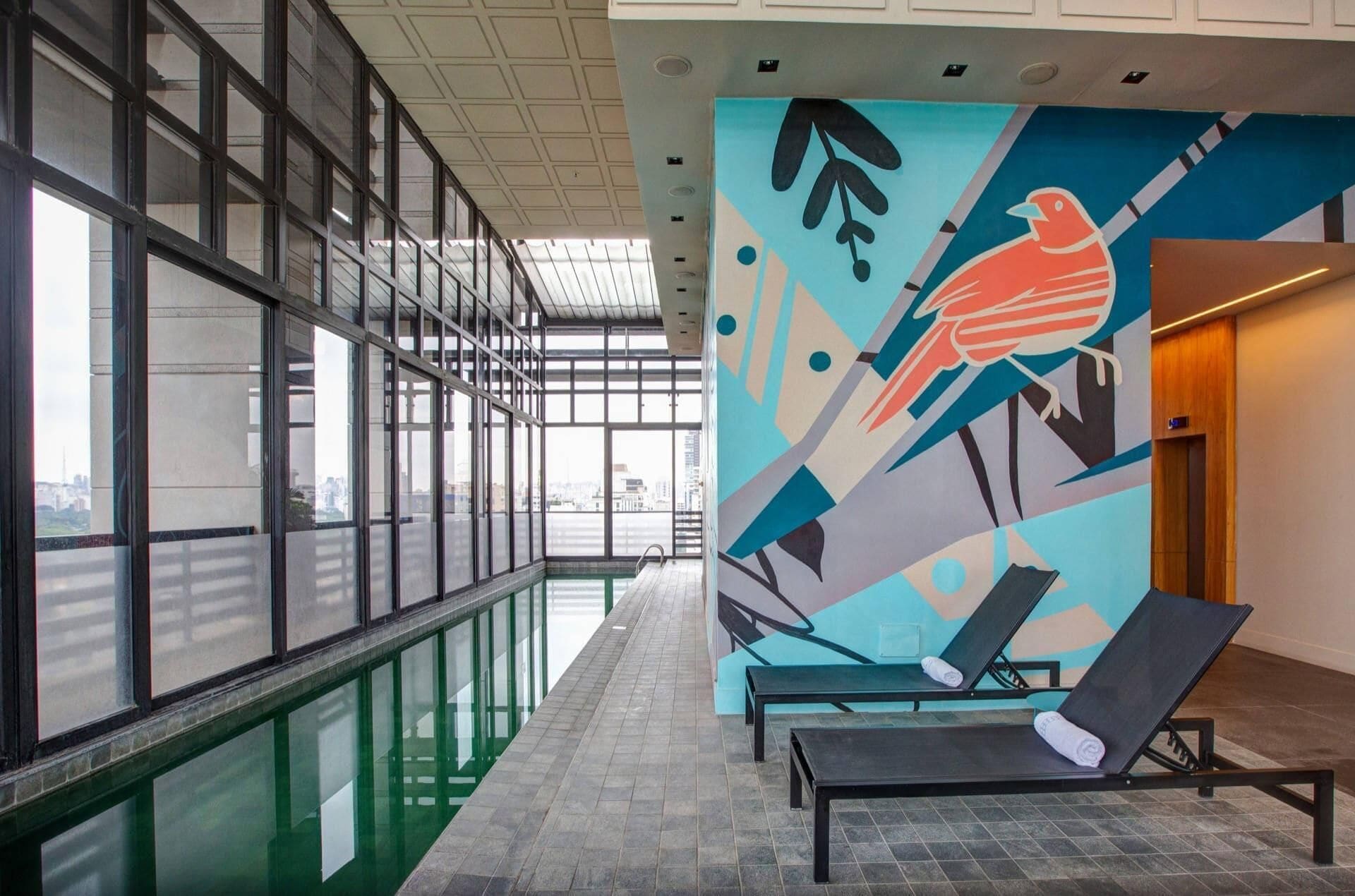 A corridor with a glass wall, a mural with abstract art, and lounge chairs with towels.