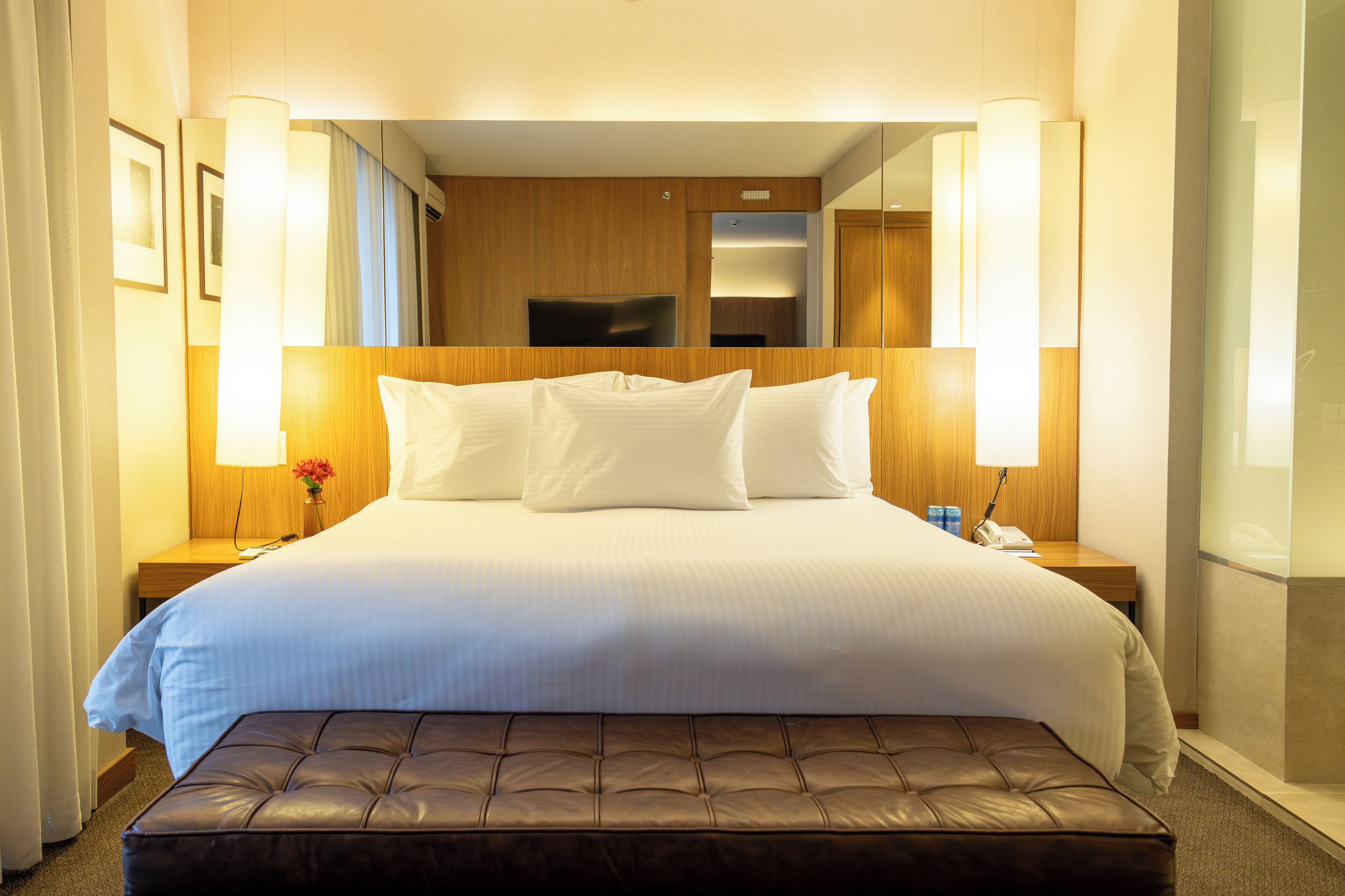 A hotel room with a large bed, a wooden headboard, a bedside table with a lamp, and a small table with a vase of flowers.