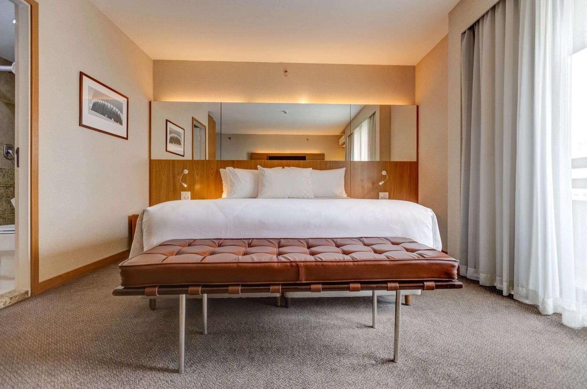 A hotel room with a large bed, a wooden headboard, a tufted bench, and a view of a cityscape through the window.