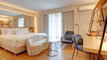 Una habitación de hotel con una cama grande, una mesita redonda pequeña, un sillón gris, un escritorio con una lámpara y un equipo de aire acondicionado.